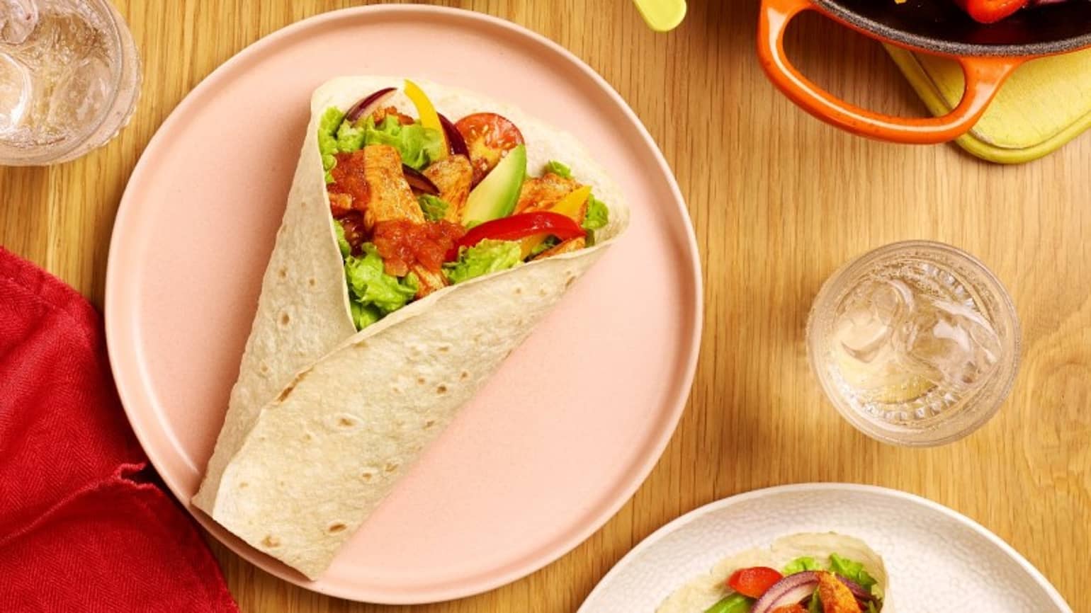 un wrap aux poulet, poivron garnis de crudité sur une assiette rose avec un verre d’eau et une poêle à proximité